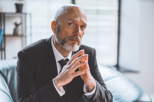 Wise senior businessman seated in office, deep in thought, gazes directly into camera. Seasoned professional with profound understanding of business world. Senior businessman's thoughtful expression. High quality photo
