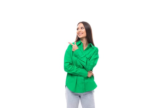 young european female model with well-groomed black hair dressed in a green shirt points her finger to the side on a white background with copy space.