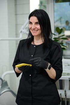 Authentic portrait of Caucasian beautiful dark-haired female doctor in black medical uniform and gloves, holding a mobile phone, smiling looking aside, standing in her newest modern dentist's office