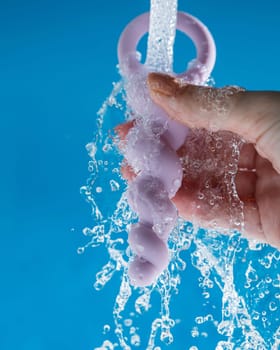 Woman holding lilac anal beads under running water on blue background. Sex toy hygiene concept