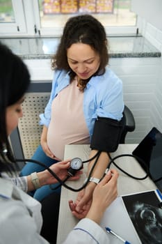 Happy pregnant woman visits gynecologist doctor in maternity hospital or medical clinic for pregnancy consultation. Obstetrician checks the blood pressure of a gravid expectant mother with a tonometer