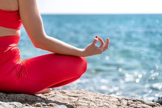 A young woman in red leggings and a red top with long loose hair practices yoga outdoors by the sea on a sunny day. Women's yoga, fitness, Pilates. The concept of a healthy lifestyle, harmony