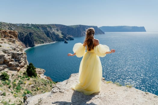 Woman in a yellow dress on the sea. Side view Young beautiful sensual woman in yellow long dress posing on a rock high above the sea at sunset. Girl in nature against the blue sky.