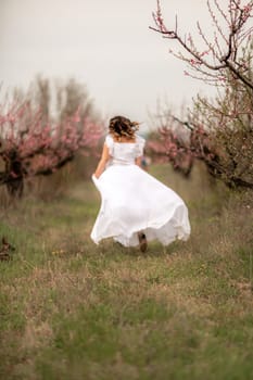 Woman peach blossom. Happy woman in white dress walking in the garden of blossoming peach trees in spring.