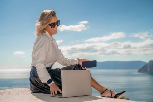 Freelance women sea working on a computer. Pretty middle aged woman with computer and phone outdoors with beautiful sea view. The concept of remote work