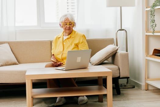 Happy elderly woman with laptop typing in headphones smile sitting at home video call, bright modern interior, lifestyle online communication. High quality photo