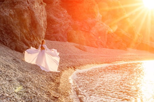 Mysterious woman silhouette long hair walks on the beach ocean water, sea nymph wind listens to the wave. Throws up a long white dress, a divine sunset. Artistic photo from the back without a face.