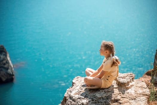 Happy girl perched atop a high rock above the sea, wearing a yellow jumpsuit and braided hair, signifying the concept of summer vacation at the beach