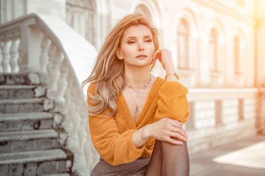 A middle-aged woman looks like a good blonde with curly beautiful hair and makeup on the background of the building. She is wearing a yellow sweater