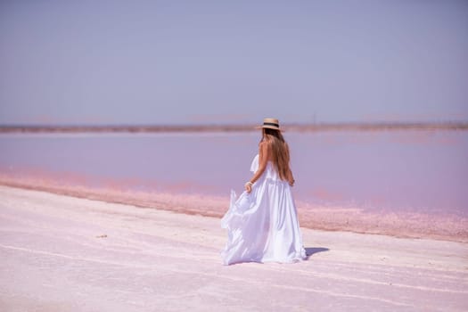 Woman in pink salt lake. She in a white dress and hat enjoys the scenic view of a pink salt lake as she walks along the white, salty shore, creating a lasting memory