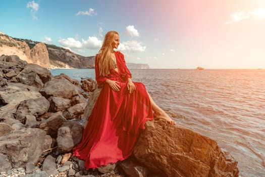 Red dress sea woman. A blonde with flowing hair in a long red dress sits on a rock near the sea. The concept of trips, a photo shoot at the sea.