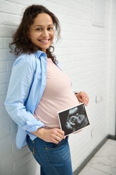 Happy adult pregnant woman with big belly, smiling looking at camera, holding the ultrasound scan of her baby in womb. Pregnancy 30 week. Obstetrics and gynecology concept. Health care