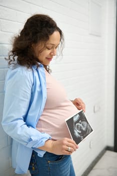 Confident multi ethnic adult pregnant woman holding the ultrasonography of her baby, standing against white brick wall background. The concept of screening in the 1st and 2nd trimester of pregnancy