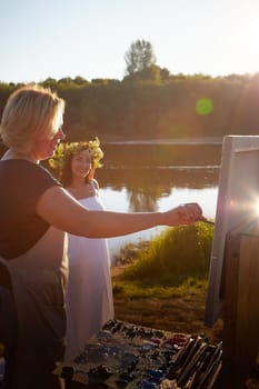 Adult female artist painting picture near water of river or lake in nature. Girl in white sundress and flower wreath for holiday of Ivan Kupala in nature at sunset near artist