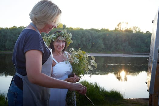 Adult female artist painting picture near water of river or lake in nature. Girl in white sundress and flower wreath for holiday of Ivan Kupala in nature at sunset near artist