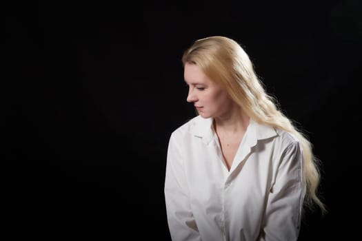 Young beautiful blonde girl in white shirt posing on a black background. Adult woman model posing alone in dark studio