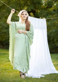 Pretty pregnant woman with big belly stands near clothesline with bed sheet, young girl takes care of sensitive skin's microbiome, holds hand on tummy outside in sunny day, looking away.Vertical plane