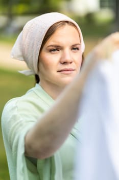 Portrait of young pretty white woman hanging up laundry on clothes line outside, drying bed linens. Safe laundry detergents, organic washing gel, fabric softener concept. Skin health care. Vertical