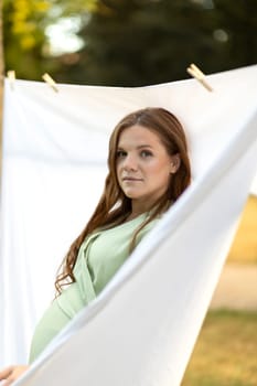 Pretty white pregnant woman hanging up laundry on clothes line outside, drying bed linens, bedding. Safe laundry detergents, organic washing gel, fabric softener concept. Skin,health care. Vertical