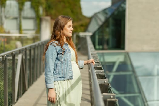 Beautiful white pregnant woman standing near modern eco green high tech building. Young female wears casual clothes. Futuristic eco-friendly estate,green district city architecture Horizontal plane