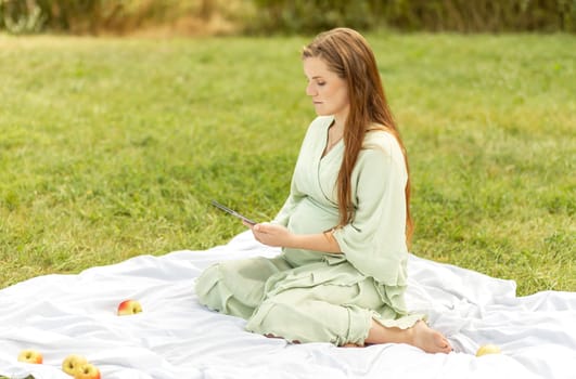 Young pretty white pregnant girl holding, reading something on tablet,gadget,resting in park,sitting on cover,blanket in park,meadow. Healthy walk outdoors. Preparation for childbirth. Horizontal