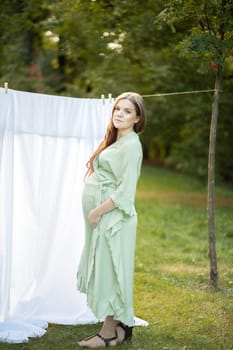 Beautiful pregnant woman with big belly stands near clothesline with bed sheet, young female takes care of sensitive skin's microbiome, drying cloth in fresh air outside. Vertical plane