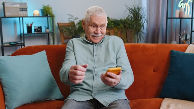 Happy senior grandfather man use mobile smartphone typing browsing say Wow yes found out great big win good news celebrate lottery jackpot, victory gesture. Elderly guy at home in room sitting on sofa