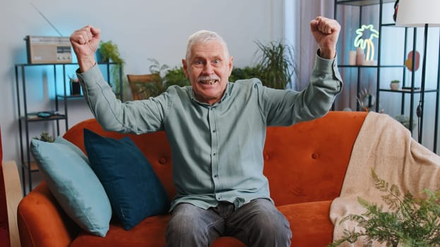 Happy senior grandfather man shouting, celebrating success, winning, goal achievement good news raising fists in gesture I did it, lottery luck at modern home apartment. Elderly guy in room on couch