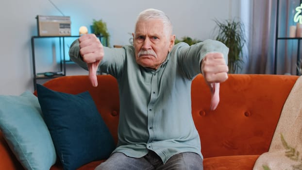 Dislike. Upset senior grandfather man showing thumbs down sign gesture, expressing discontent disapproval dissatisfied bad work at modern home apartment indoors. Displeased elderly guy in room on sofa