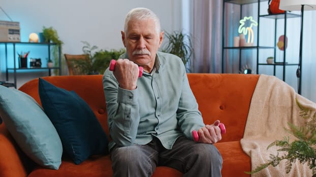 Senior grandfather man doing dumbbells weight lifting fitness exercises at home apartment indoors. Elderly old guy workout training strength practicing effort in living room on couch. Sport motivation