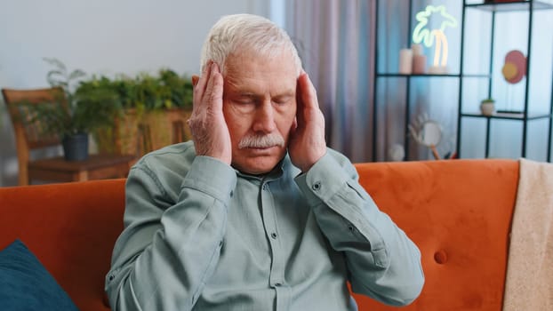 Displeased senior grandfather man rubbing temples to cure headache problem, suffering from tension and migraine, stress grimacing in pain high blood pressure at home. Elderly guy on couch. Lifestyles