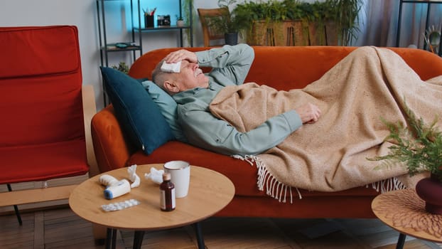 Grandfather ill man suffering from cold or allergy lying on sofa in room at home apartment. Sick senior guy with towel on head blows sneezes wipes snot into napkin. Coronavirus quarantine, pandemic