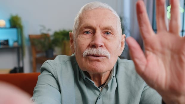 POV of senior grandfather man making online selfie video call talking waving hand looking at camera at home apartment indoors. Communication, meeting. Elderly blogger making smartphone conversation