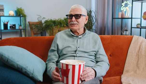 Senior grandfather man in 3D glasses on couch eating popcorn snacks and watching interesting TV serial sport game, film online social media movie content at home apartment. Guy enjoying entertainment