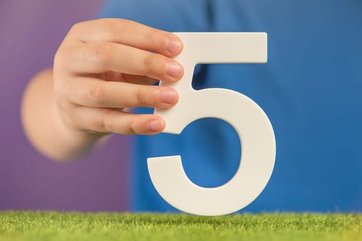Number 5 in hand. A hand holds a white number five on green grass and a purple background, five percent per annum. Concept of victory day in the Netherlands and Mexico, 5 May