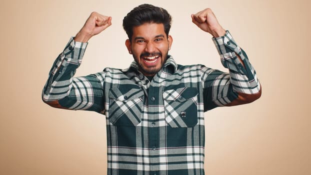 Happy joyful indian man shouting, raising fists in gesture I did it, celebrating success, winning, birthday, lottery jackpot goal achievemen, good news. Young hindu guy on beige studio wall background