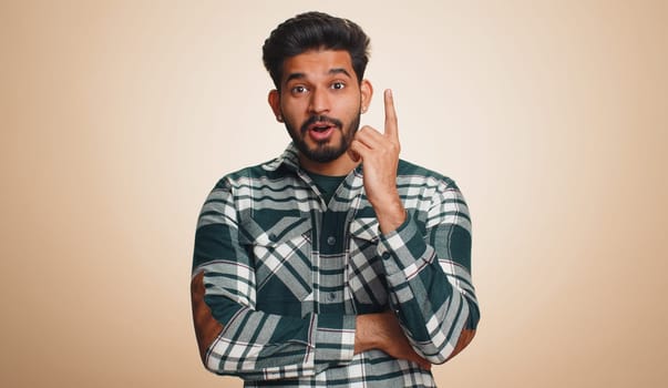 Eureka. Inspired young indian man pointing finger up with open mouth, having good idea, plan, startup, showing inspiration motivation gesture, problem solution. Bearded hindu guy on beige background