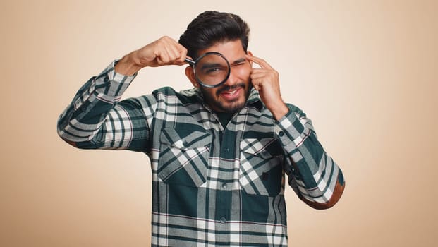 Investigator researcher scientist man holding magnifying glass near face, looking into camera with big zoomed funny eyes, searching, analysing. Handsome indian guy isolated on beige studio background