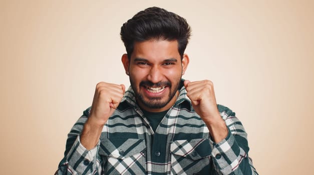 Happy joyful indian man shouting, raising fists in gesture I did it, celebrating success, winning, birthday, lottery jackpot goal achievemen, good news. Young hindu guy on beige studio wall background