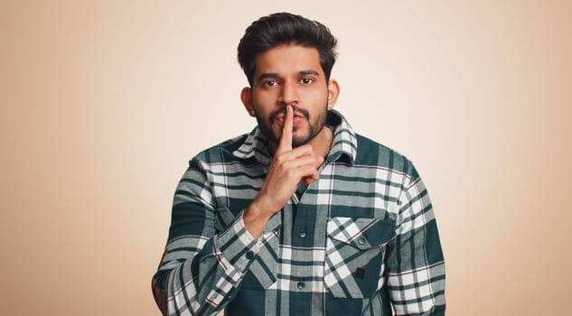 Shh be quiet please. Young indian man presses index finger to lips makes silence gesture sign do not tells secret, stop gossip. Handsome bearded hindu guy isolated alone on beige studio background
