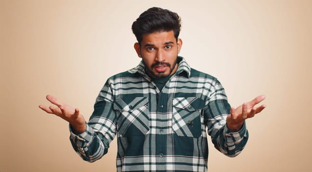 What Why. Sincere irritated indian man raising hands in indignant expression asking reason of failure demonstrating disbelief irritation by trouble. Handsome confused hindu guy on beige background