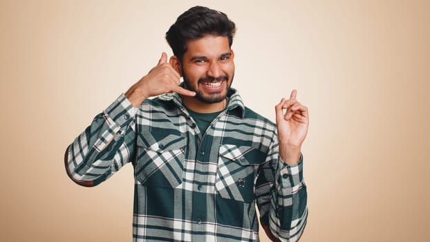 Call us, here is contact number. Handsome bearded indian man in shirt looking at camera doing phone gesture like says hey you call me back conversation. Hindu guy isolated on beige background, indoors