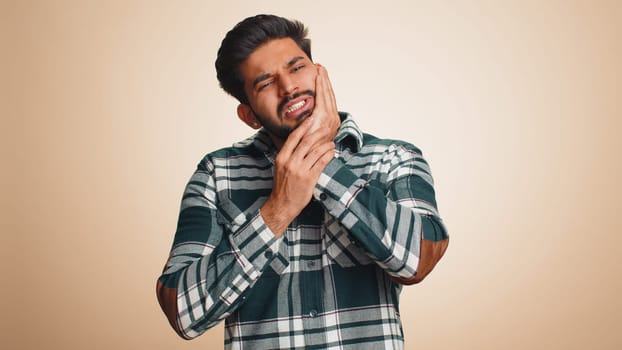 Indian bearded man touching sore cheek suffering from toothache cavities or gingivitis waiting for dentist appointment gums disease. Handsome hindu guy indoors studio shot isolated on beige background