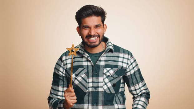 Magician wizard man gesturing with magic wand fairy stick, making wish come true, casting magician spell, advertising holidays sale discount. Handsome indian guy isolated on beige studio background