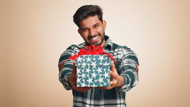 Positive smiling young indian man presenting birthday gift box stretches out hands, offer wrapped present career bonus, celebrating party, sale. Bearded hindu guy isolated on beige studio background