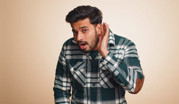 I cant hear you. What. Young indian man trying hear, looking confused, frowning, keeping arm near ear for louder voice, asking to repeat, to hear information, deafness. Hindu guy on beige background