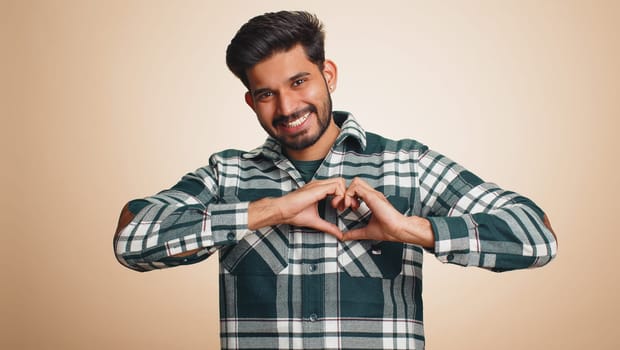 Man in love. Smiling bearded indian man 25 years old makes heart gesture demonstrates love sign expresses good feelings and sympathy. Handsome hindu young guy isolated alone on beige studio background