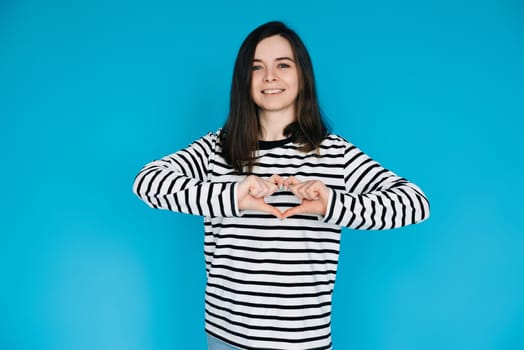 Joyful Woman in Striped Sweater Smiling, Gesturing Heart Hands - Symbol of Love and Happiness - Isolated on Blue Background - Perfect for Positive Emotions, Love, and Lifestyle Concepts.