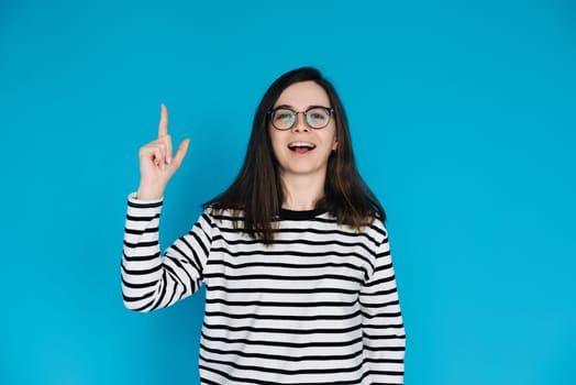 Excited Cheerful Woman in Striped Sweater with Glasses Pointing Up - Energetic Female Model Gesturing towards Empty Space - Isolated on Blue Background - Perfect for Advertising, Promotions Concepts