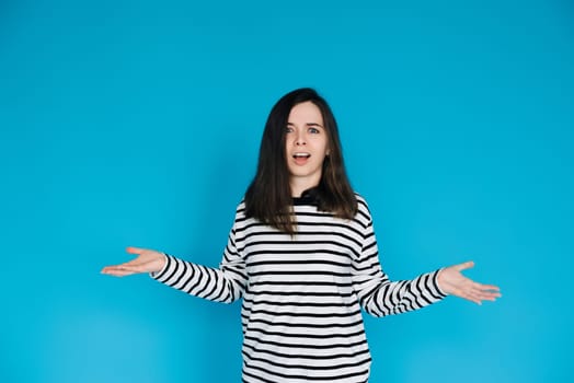 Captivating Portrait of Astonished Woman in Striped Pullover - Surprised and Amazed Female Model with Arms Extended - Isolated on Blue Background - Perfect for Expressing Shock, Surprise, and Emotion.